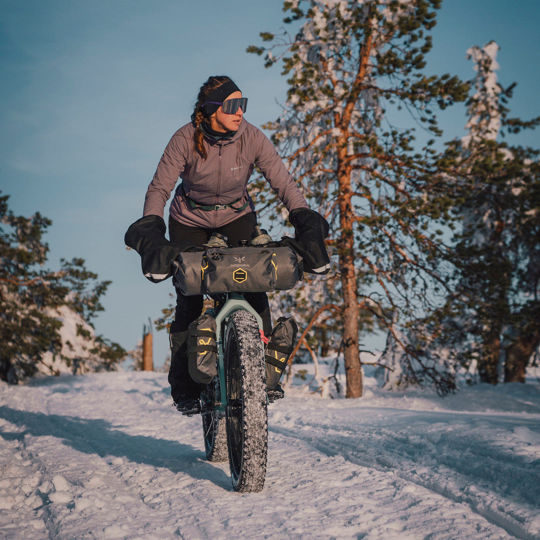 A person riding a fat-tire bicycle on a snowy trail in a forest, wearing a taupe-gray long-sleeve top, black pants, gloves, and goggles, with a dark bike bag carrying a yellow logo.