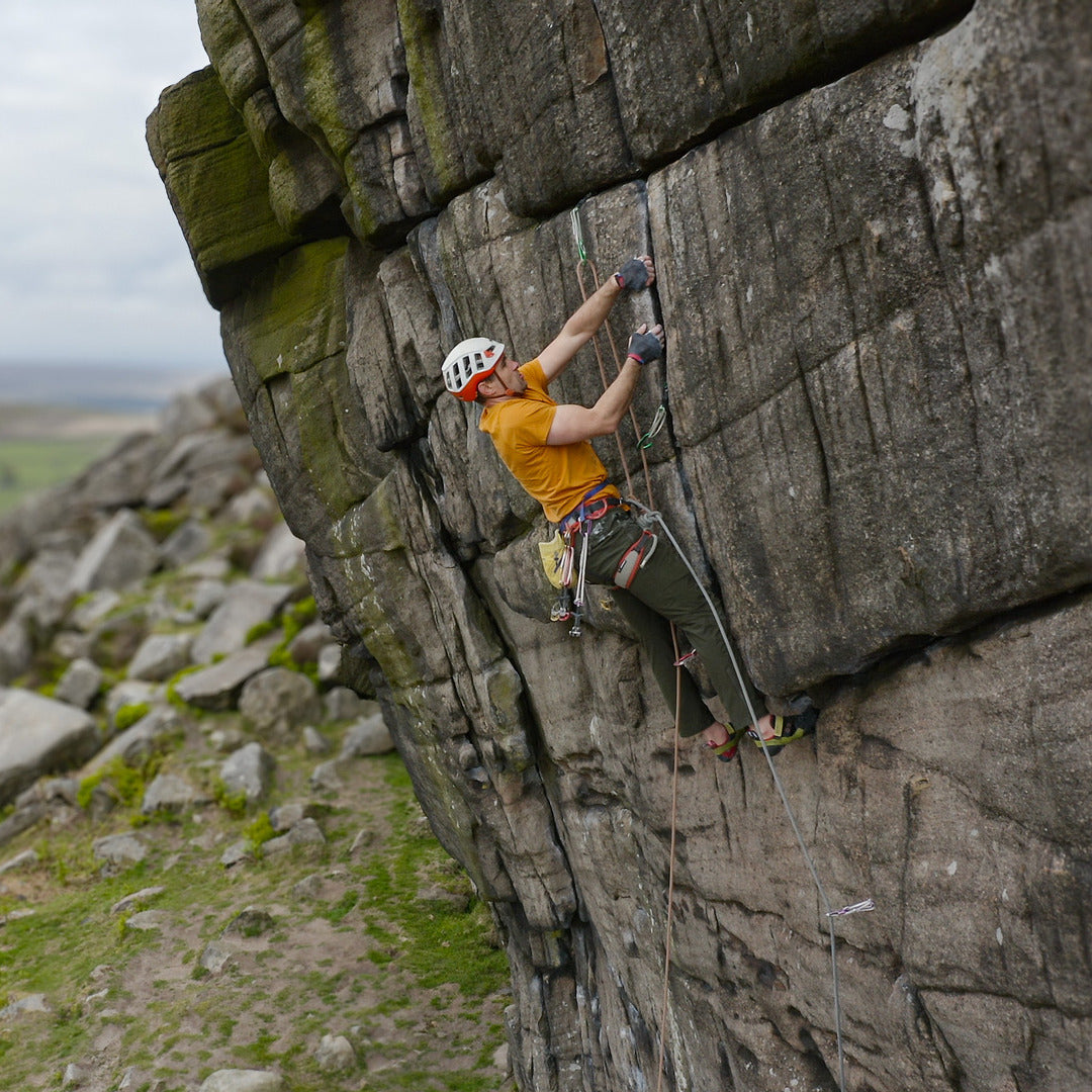 A rock climber wearing a white helmet, orange shirt, and green pants ascends a steep gray stone cliff using handholds and ropes, with rocky terrain and grass below.