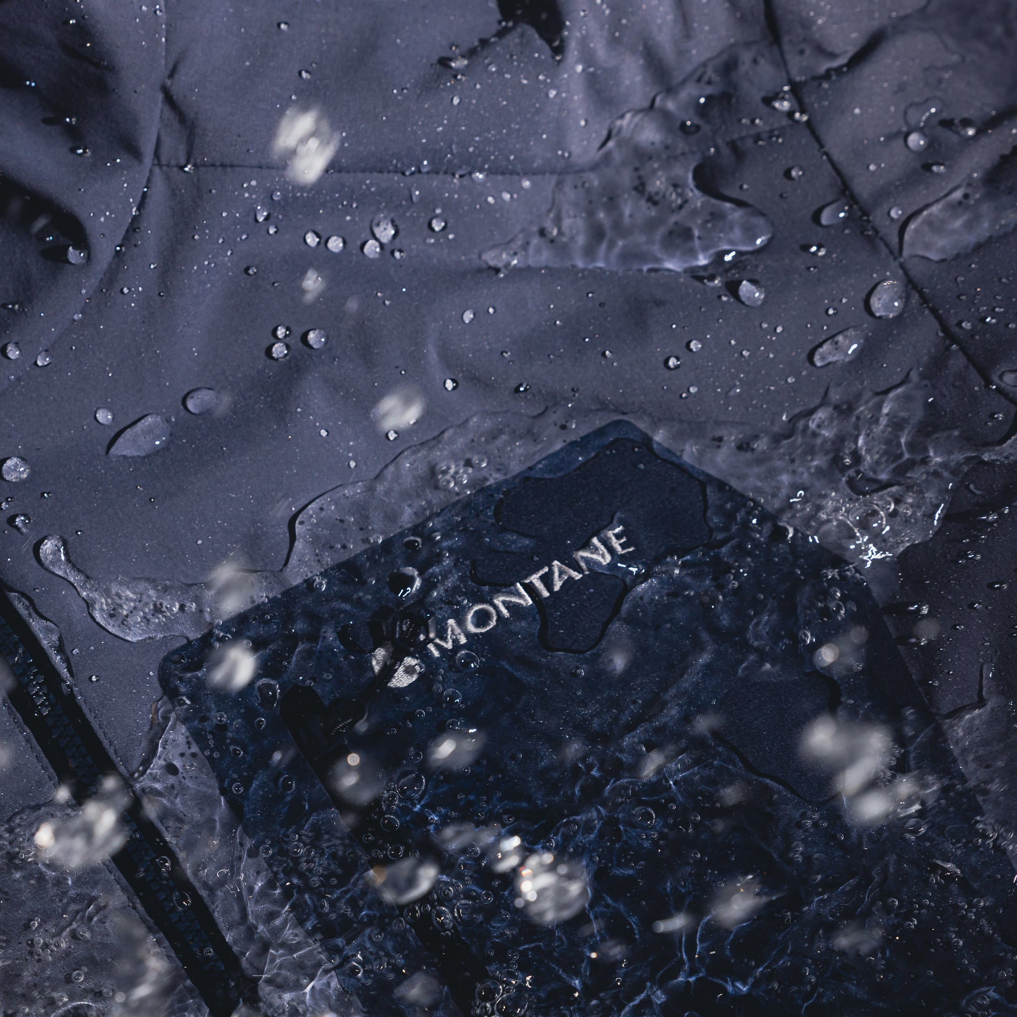 A close-up of a dark navy blue waterproof jacket covered in water droplets, with a matching Montane-branded pouch resting on the wet fabric.