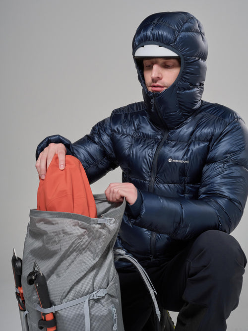 Person in a blue puffer jacket with a hood, holding an orange item, against a gray background.